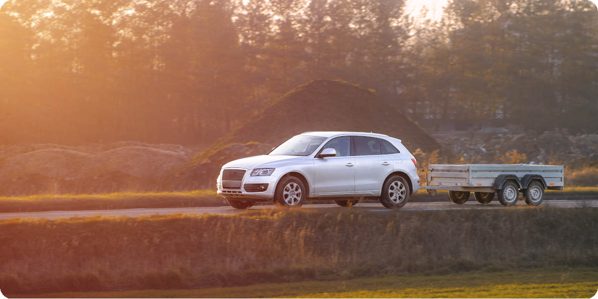 Ein silbernes Auto zieht einen Anhänger auf einer Landstraße im Sonnenuntergang.