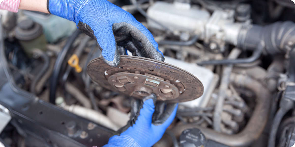 Zwei Hände mit blauen Gummihandschuhen halten eine verschlissene Kupplungsscheibe über dem Motorblock.